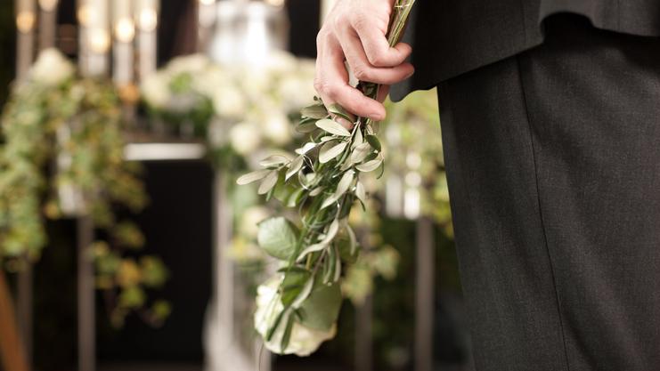 Mann in schwarzem Anzug hält einen Strauß mit grünen Blättern und einer weißen Rose vor einem dekorierten Hintergrund.