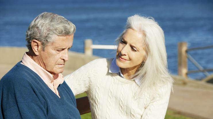 Ein älteres Paar sitzt auf einer Bank am Wasser, die Frau hält den Blick des Mannes sanft und einfühlsam.